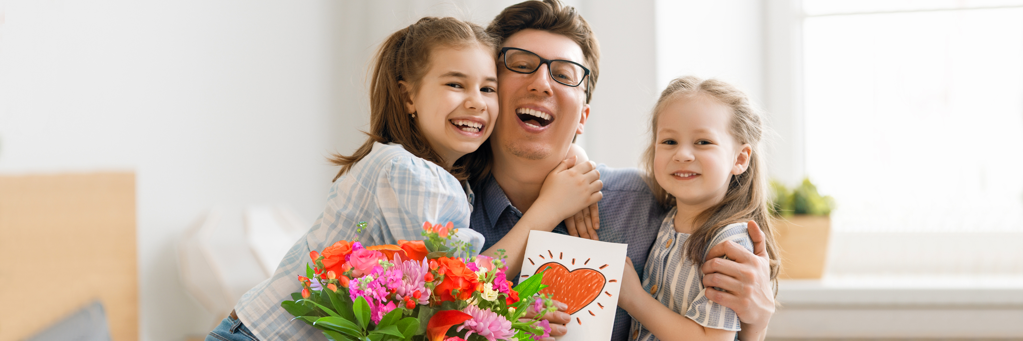 A joyful father embraces his two daughters, surrounded by colorful flowers and a heartfelt drawing.