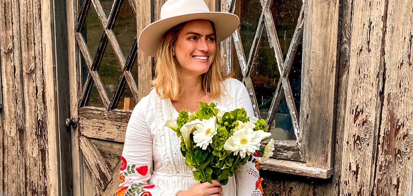 A woman in a stylish hat holding a bouquet of white flowers, standing by a rustic wooden door.