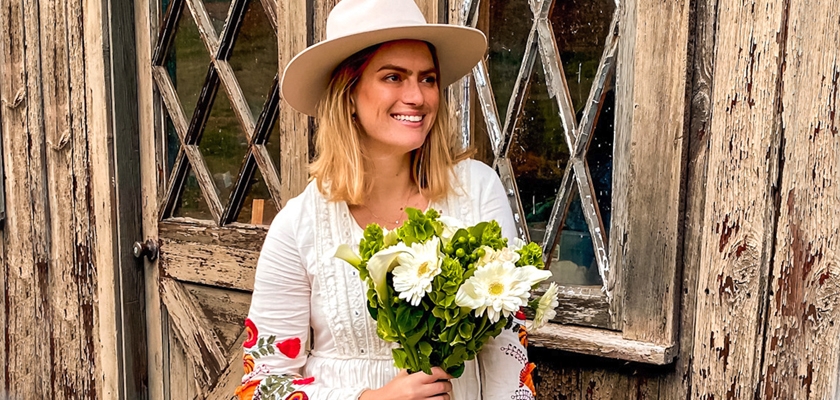 A woman in a stylish hat holding a bouquet of white flowers, standing by a rustic wooden door.