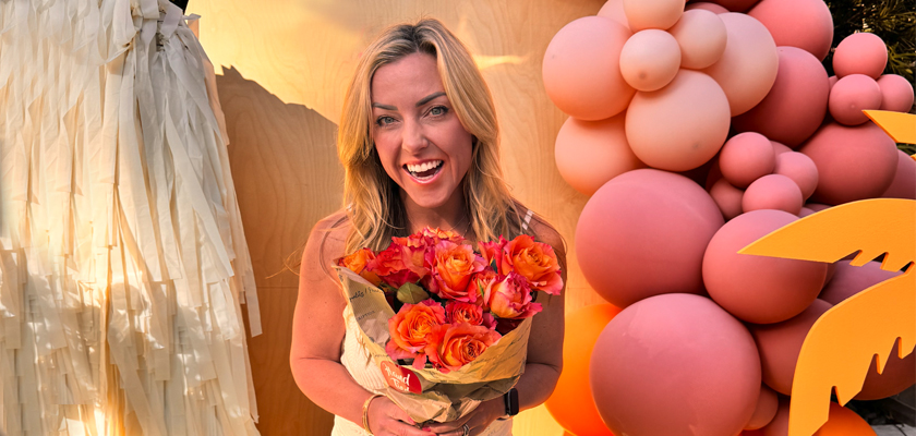 A smiling woman holds a vibrant bouquet of orange roses, surrounded by colorful balloon decor.