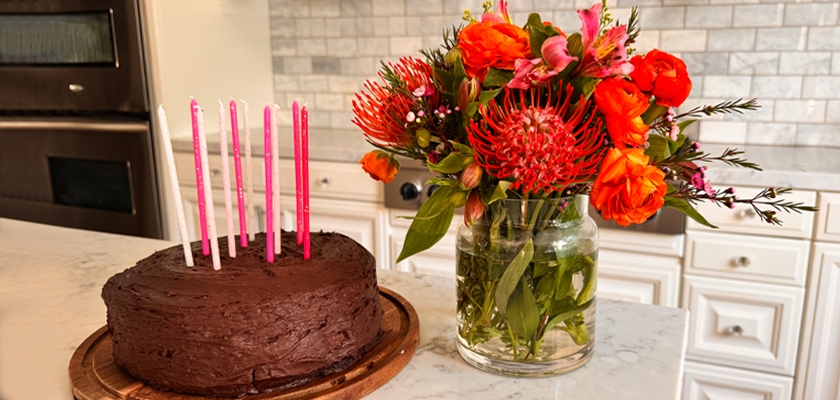 A delicious chocolate birthday cake surrounded by a vibrant floral arrangement.