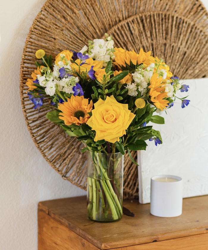 Bright floral arrangement featuring yellow roses, sunflowers, and blue blossoms in a clear vase.