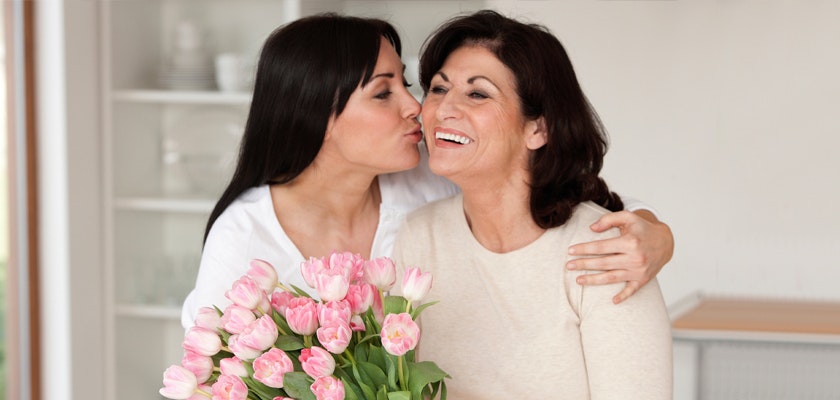 A happy woman kisses her mother while holding a bouquet of pink tulips, celebrating love and joy.