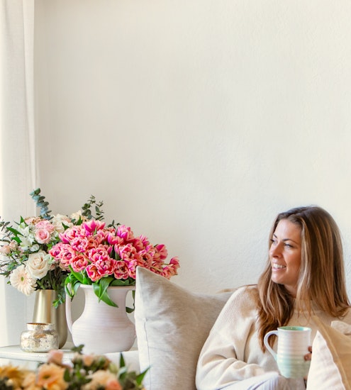 A woman enjoys a cozy moment with a warm drink, surrounded by vibrant flower arrangements.