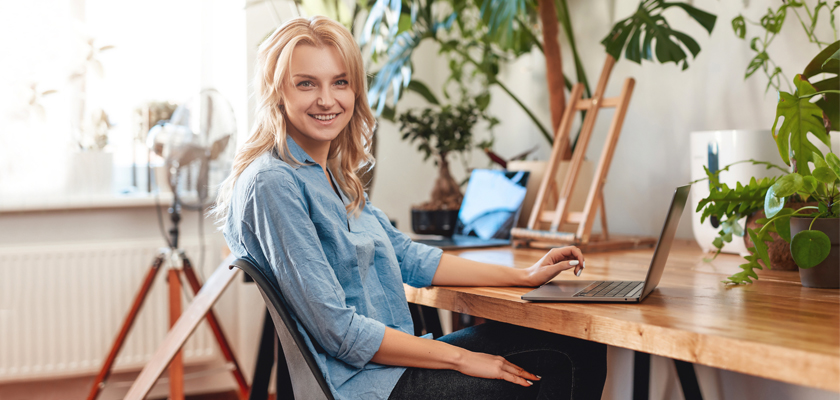 A young woman smiling while using a laptop at a cozy, green workspace decorated with plants.