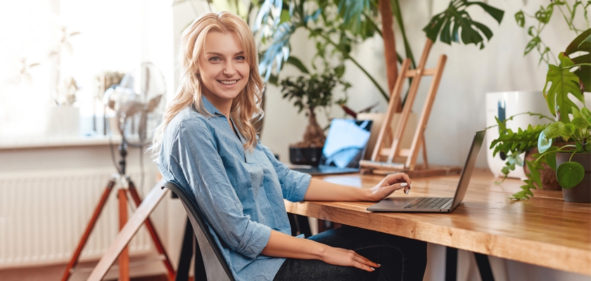 A young woman smiling while using a laptop at a cozy, green workspace decorated with plants.