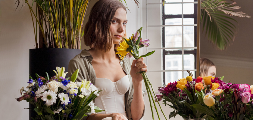 A woman holding a yellow flower, surrounded by vibrant floral arrangements in a bright studio.