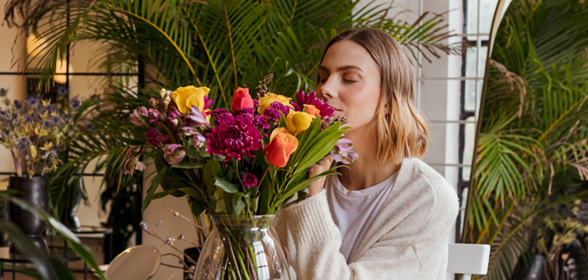 A woman enjoys a colorful bouquet of flowers, surrounded by lush greenery in a cozy indoor setting.