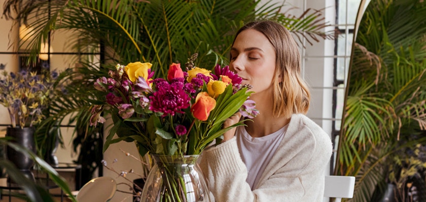 A woman enjoys a colorful bouquet of flowers, surrounded by lush greenery in a cozy indoor setting.