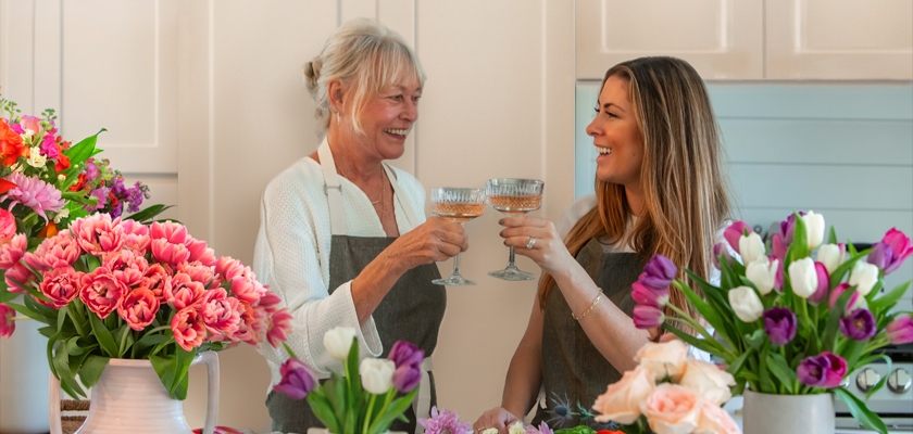 Two women toast with champagne in a bright floral workshop surrounded by vibrant bouquets.