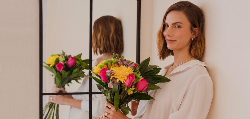 A woman holding a vibrant bouquet of pink roses and bright flowers, standing in front of a mirror.