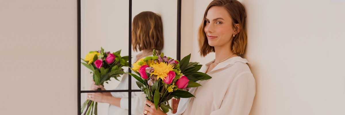 A cheerful woman holding a vibrant bouquet of flowers, standing beside a mirror.