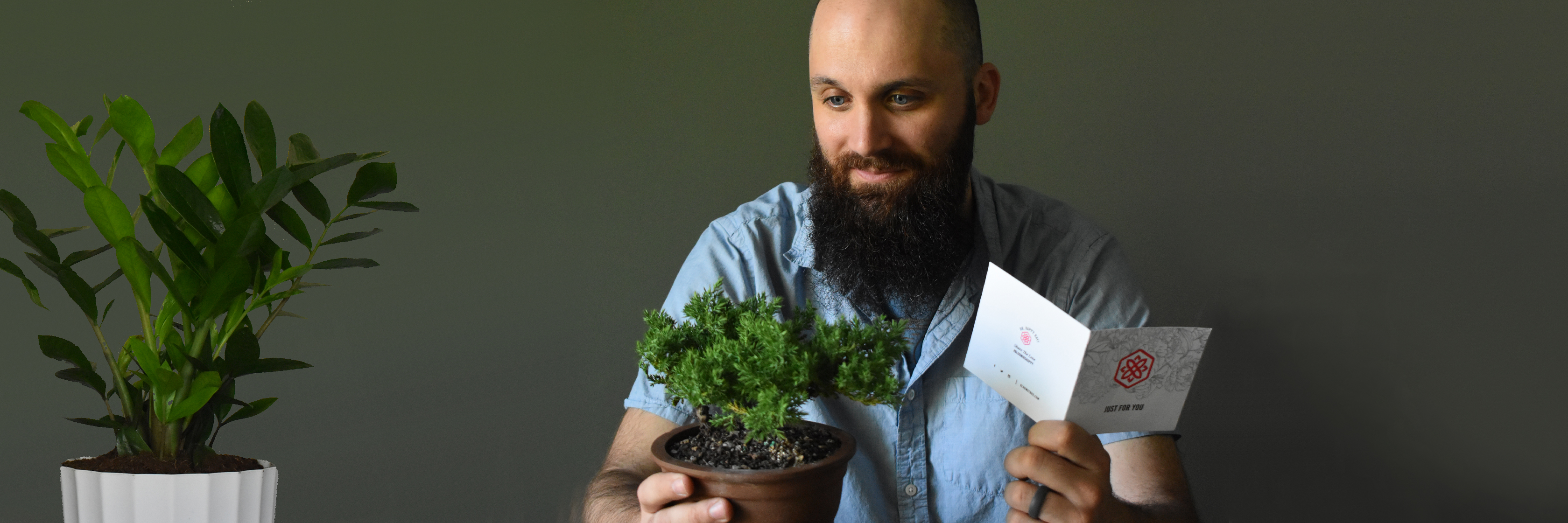 A man with a beard holding a small potted plant and a greeting card, surrounded by greenery.