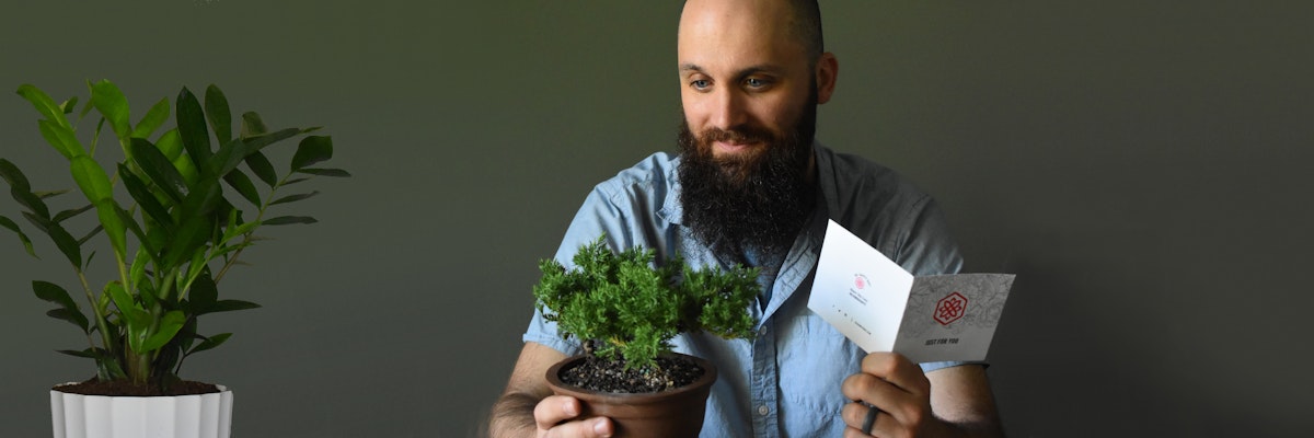 A man with a beard holding a small potted plant and a greeting card, surrounded by greenery.