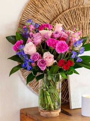 Vibrant flower arrangement featuring pink and purple roses, cheerful carnations, and greenery.