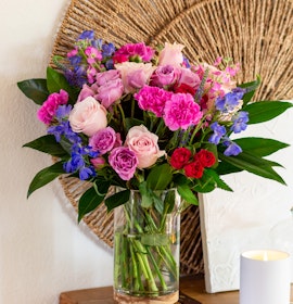 Vibrant flower arrangement featuring pink and purple roses, cheerful carnations, and greenery.