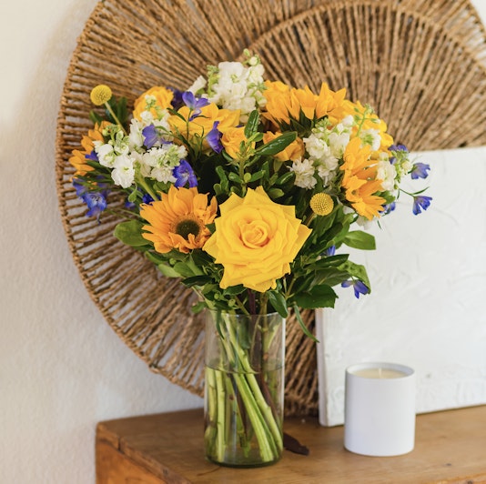 Bright bouquet of yellow roses, sunflowers, and blue flowers in a stylish glass vase.