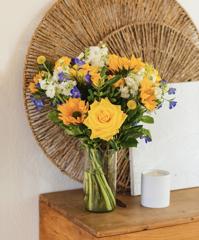 Bright bouquet of yellow roses, sunflowers, and blue flowers in a stylish glass vase.