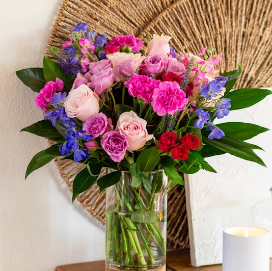 Vibrant vase filled with pink and purple roses, carnations, and blue delphiniums on a wooden table.