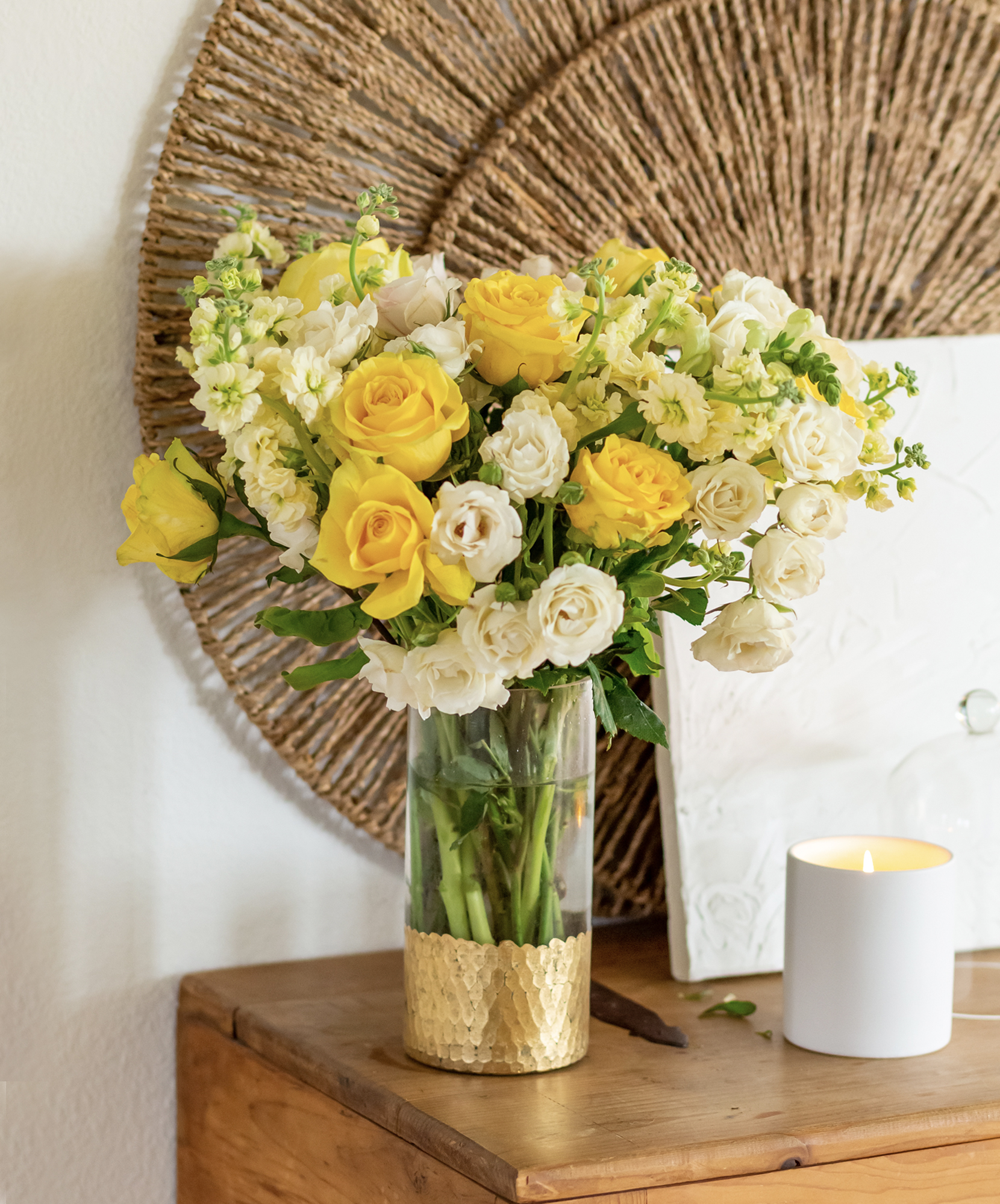 A vibrant bouquet of yellow roses and white flowers in a glass vase, complemented by a gold base.