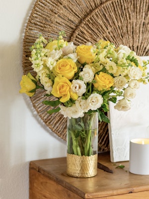 A vibrant bouquet of yellow roses and white flowers in a glass vase, complemented by a gold base.