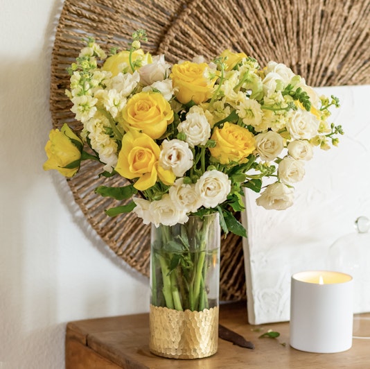 A vibrant bouquet of yellow roses and white flowers in a glass vase, complemented by a gold base.