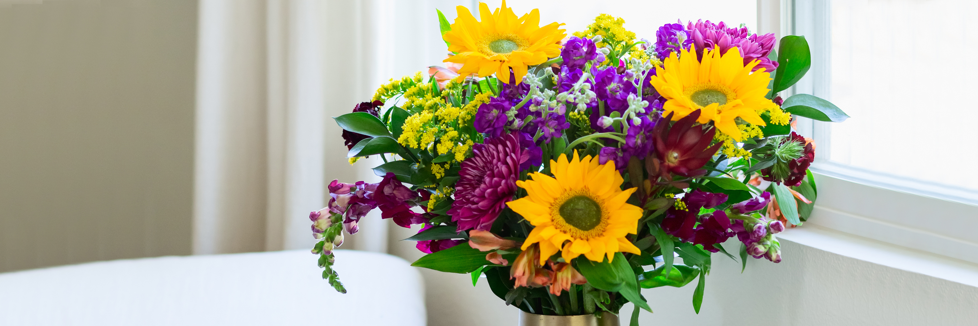 Bright and cheerful flower bouquet featuring sunflowers, purple blooms, and greenery by a window.