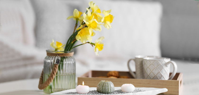 Bright yellow daffodils in a glass vase, accompanied by candles and a cozy tea setting.