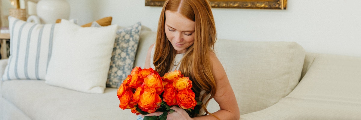 Woman with long red hair holding a vibrant bouquet of orange roses, seated on a cozy sofa.