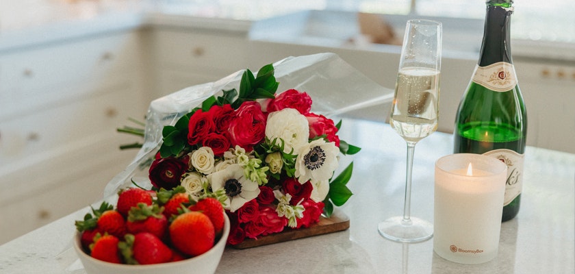 A vibrant bouquet of mixed roses and anemones beside champagne, strawberries, and a candle.