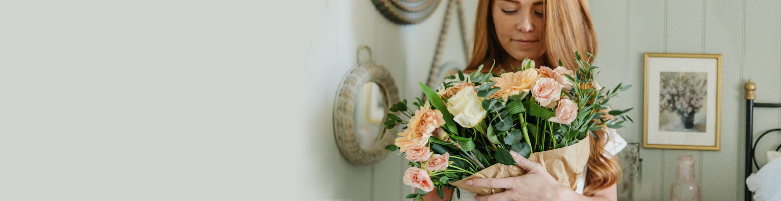 Woman holding a gorgeous bouquet of soft peach and cream flowers, surrounded by a cozy interior.