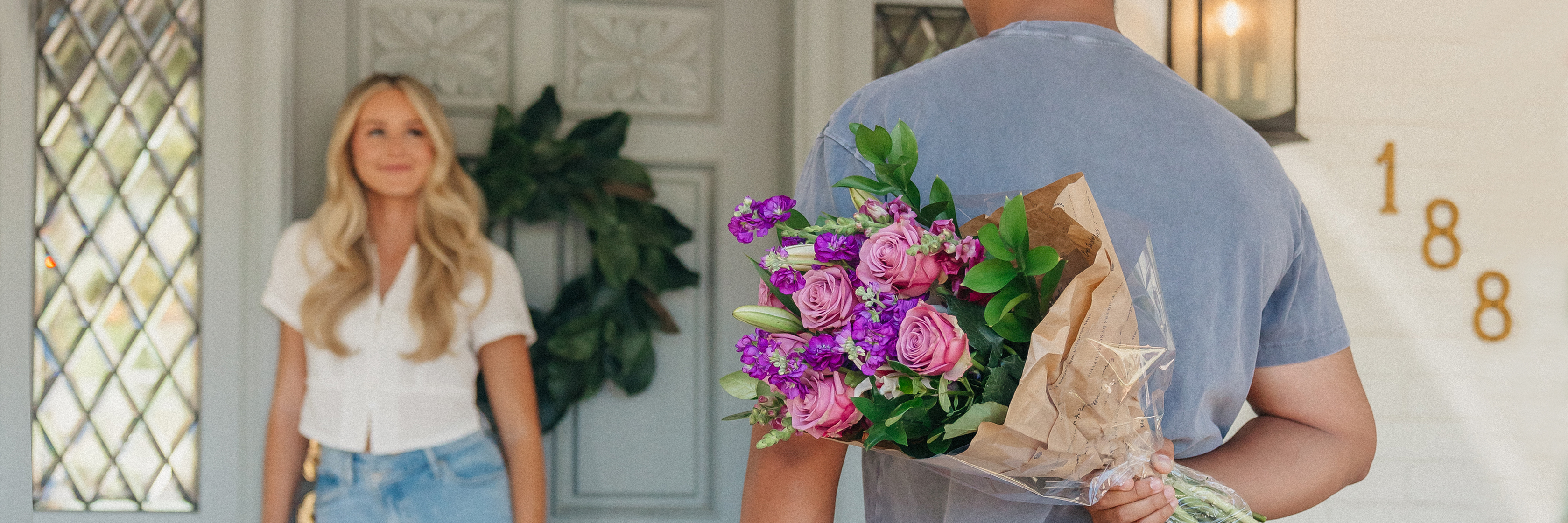 A smiling woman stands by a door as a man surprises her with a beautiful flower bouquet.