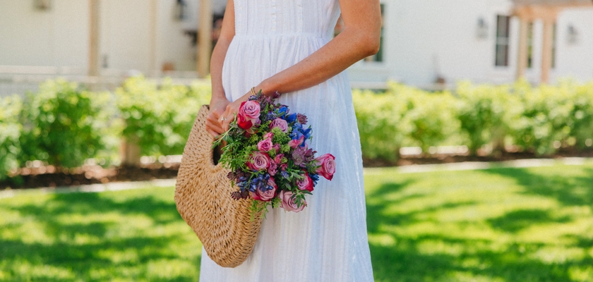 A woman in a white dress holds a woven basket filled with vibrant flowers in a lush garden.