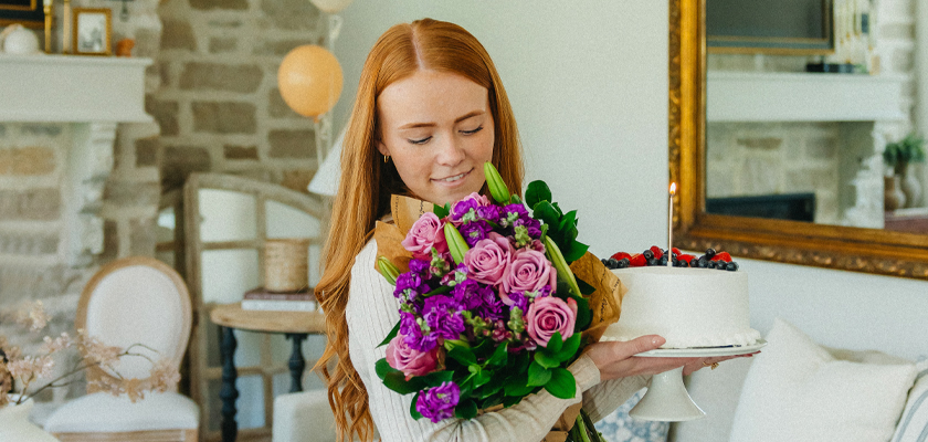 A young woman holds a colorful bouquet and a cake, celebrating a special occasion.