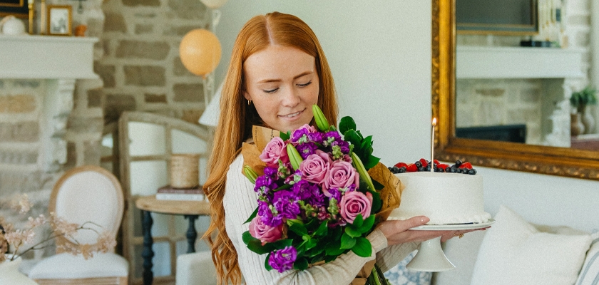 A young woman holds a colorful bouquet and a cake, celebrating a special occasion.