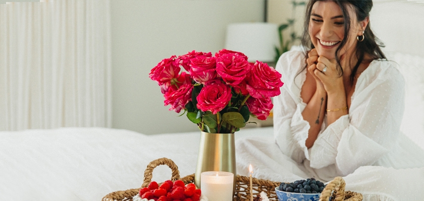 Bright pink roses in a golden vase, surrounded by fruits and a candle, creating a cheerful ambiance.