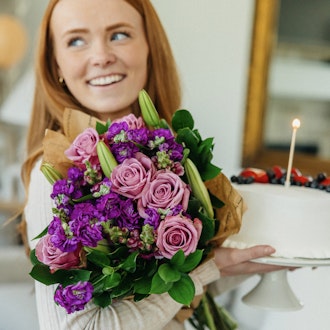 A joyful woman holds a vibrant bouquet of pink roses and purple flowers alongside a delicious cake.