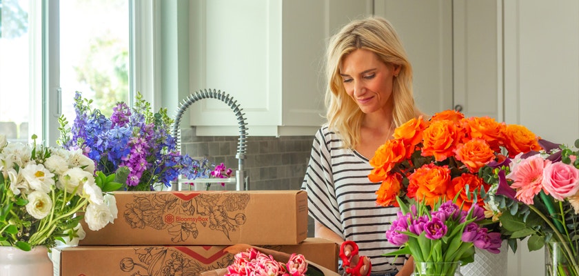 Woman arranging vibrant flowers from delivery boxes in a bright kitchen setting.