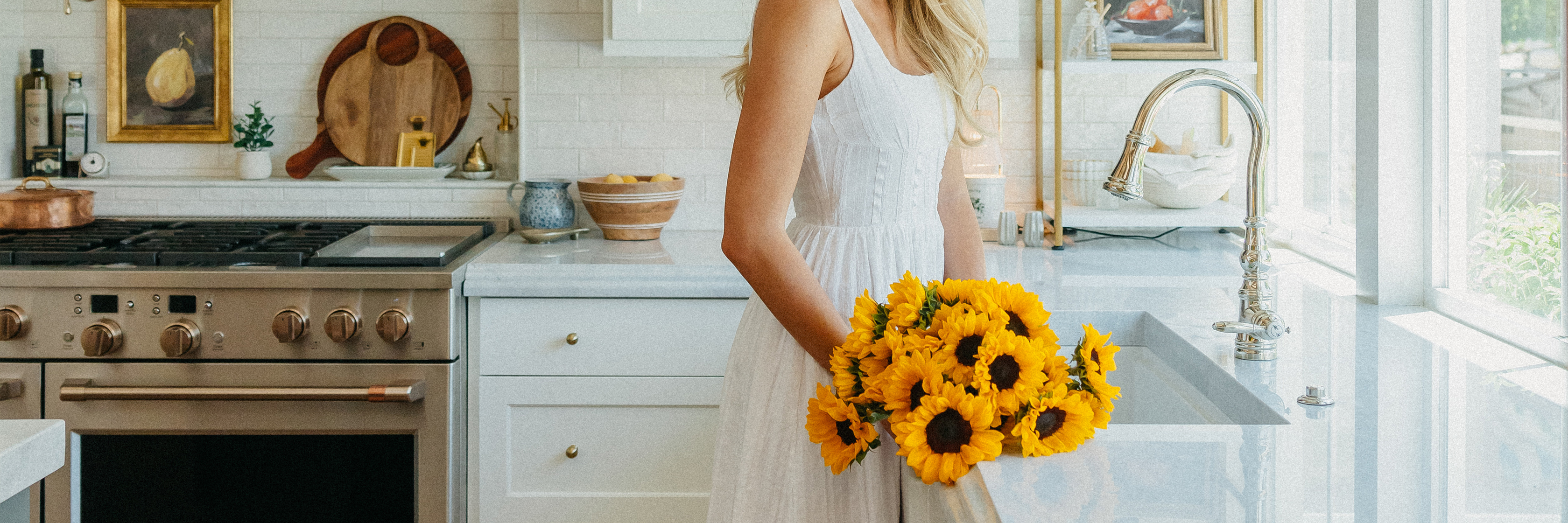 A woman in a white dress holds a vibrant bouquet of sunflowers in a bright kitchen, exuding warmth.