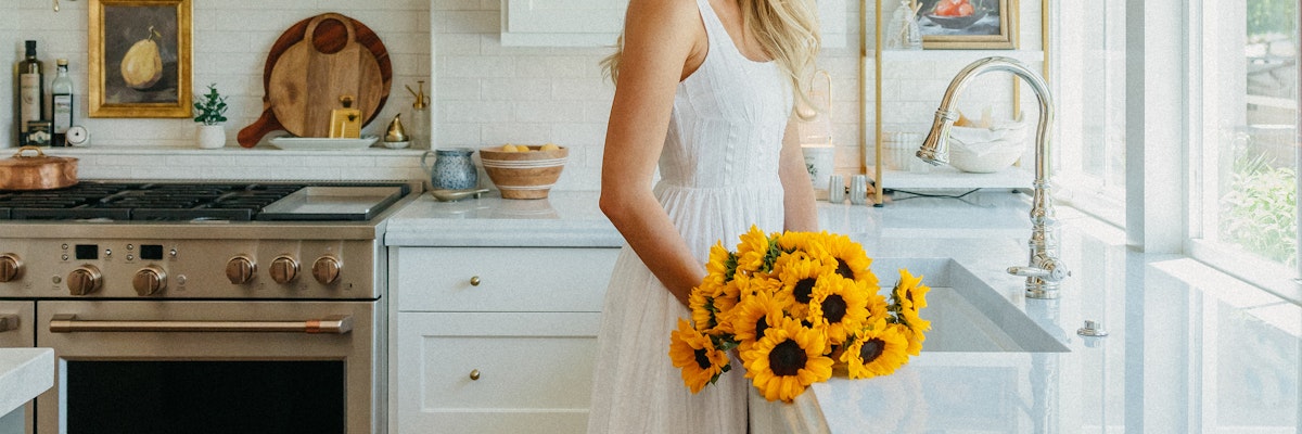A woman in a white dress holds a vibrant bouquet of sunflowers in a bright kitchen, exuding warmth.