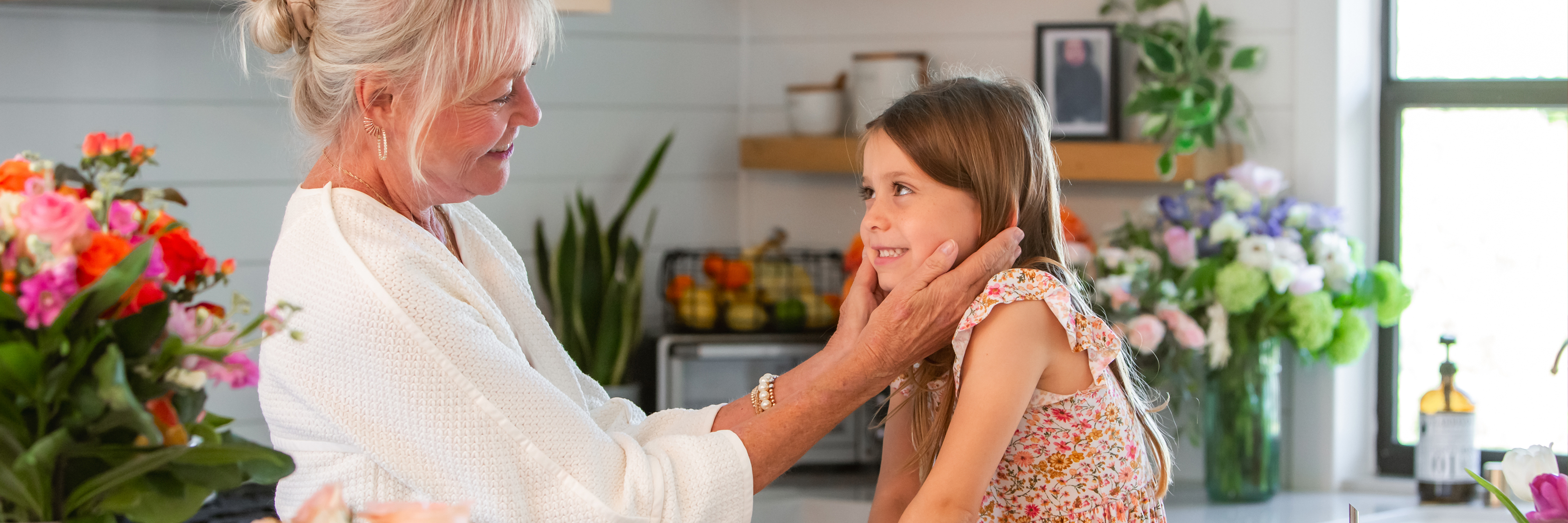 An elderly woman sharing a joyful moment with a young girl in a bright kitchen filled with flowers.
