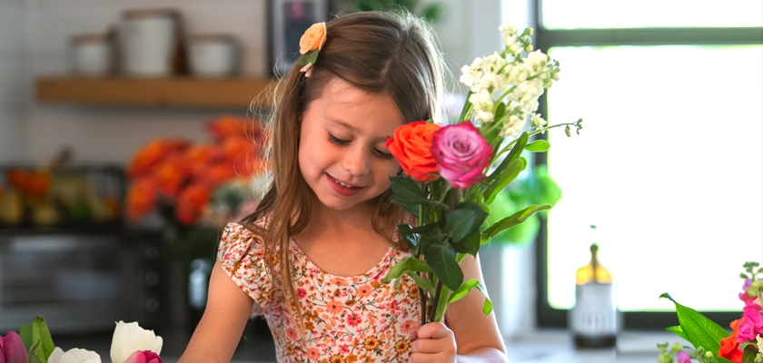 A cheerful girl in a floral dress arranging a vibrant bouquet of colorful flowers at home.