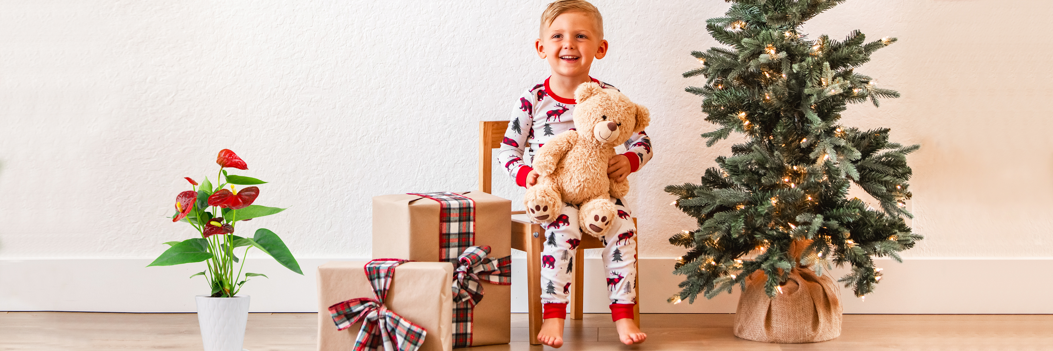 Cheerful child in festive pajamas sitting with stuffed bear by a Christmas tree and gifts.