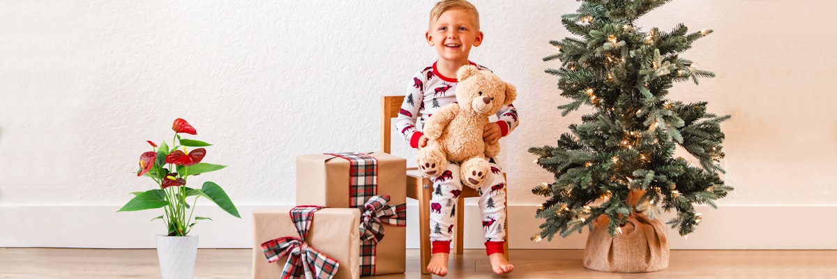 Cheerful child in festive pajamas sitting with stuffed bear by a Christmas tree and gifts.