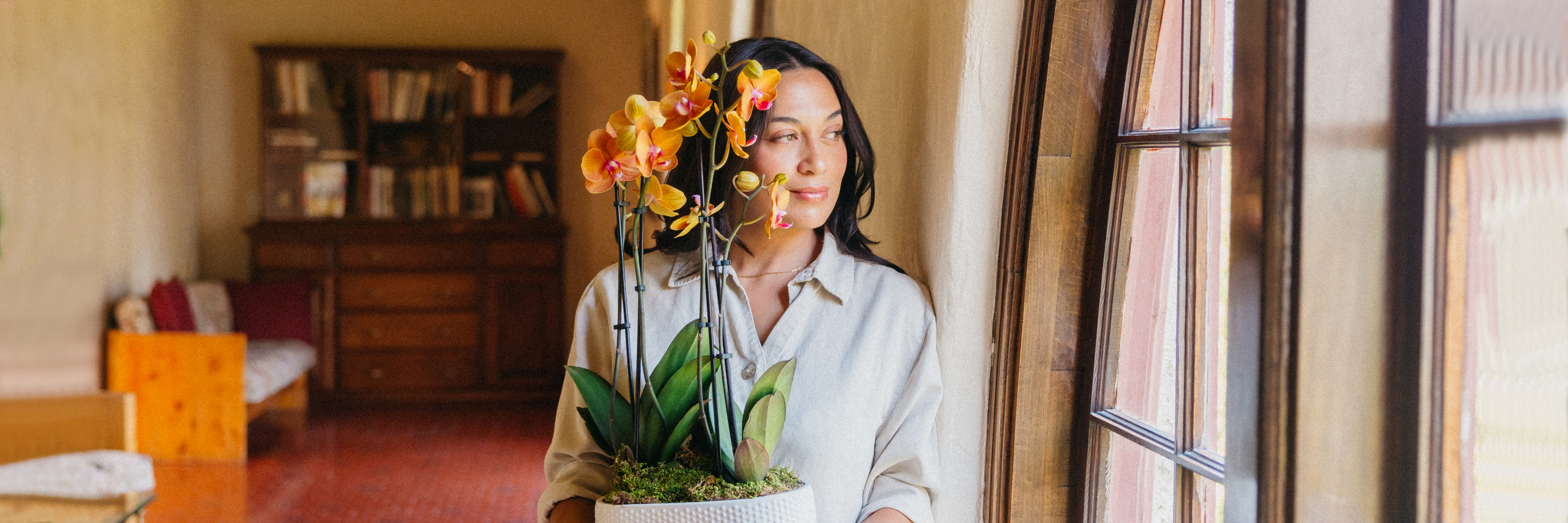 A woman holding a vibrant orchid arrangement near a sunlit window, enhancing the room's ambiance.