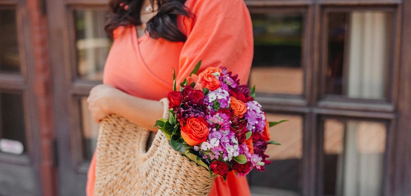 A vibrant floral bouquet featuring roses and orchids, elegantly held in a stylish woven bag.