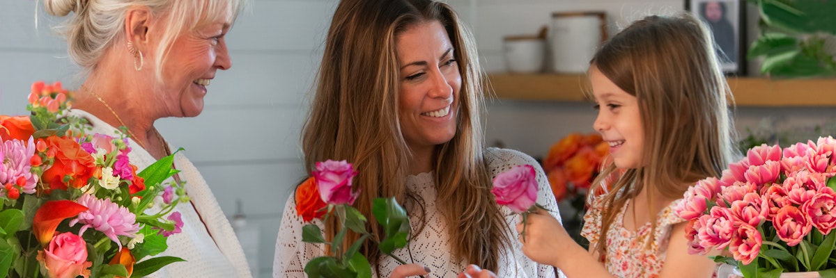 Three smiling women, including a child, share vibrant flower bouquets in a cozy indoor setting.