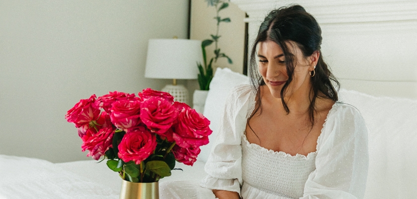 A cheerful young woman sitting on a bed beside a vibrant pink rose bouquet in a gold vase.