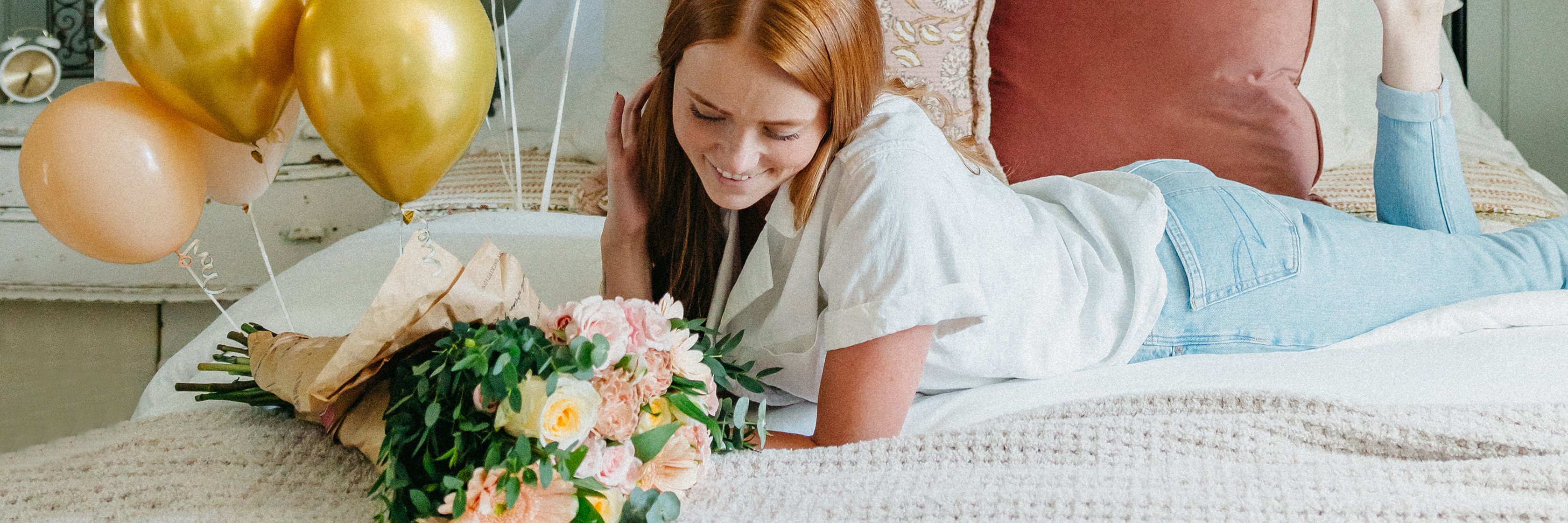 A young woman reclines on a bed, smiling with a floral bouquet and gold balloons nearby.