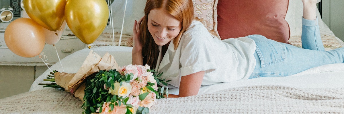 A young woman reclines on a bed, smiling with a floral bouquet and gold balloons nearby.